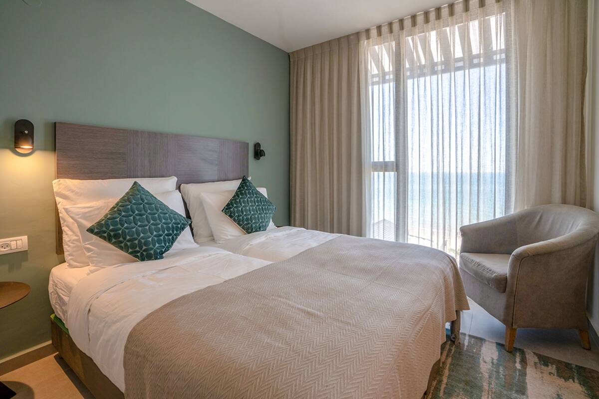 A hotel room with a large bed, two pillows, a chair, and a view of the outdoors through sheer curtains.