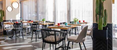 A modern dining area with tables and chairs, a cactus decoration, and a large window with sheer curtains.