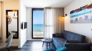 A modern living room with a large window offering a beach view, a grey sofa, a small round table, and a TV mounted on the wall. - Roxon Sea Sand Bat Yam (Bat Yam)