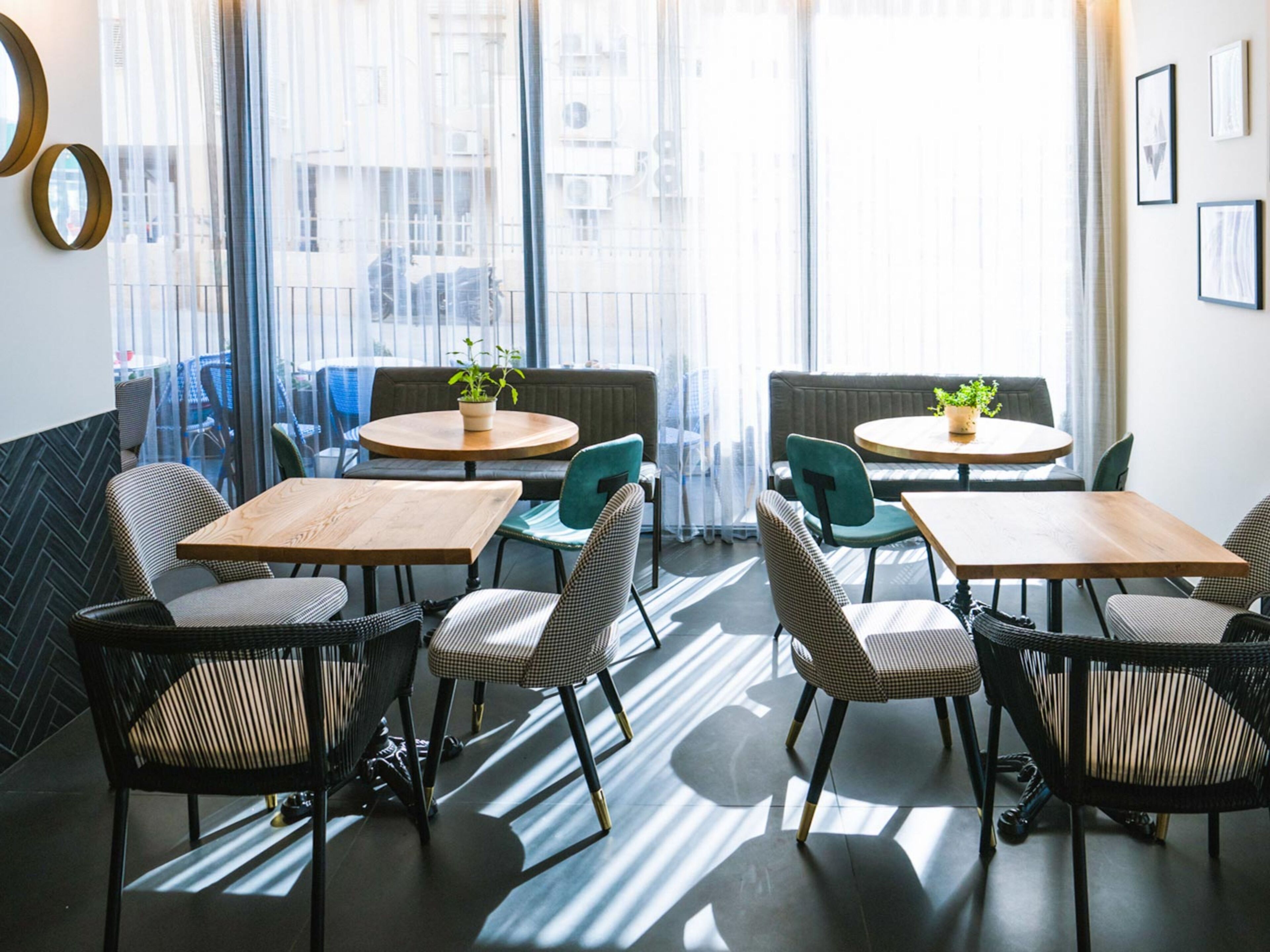 Ein modernes Café mit runden Tischen und Stühlen, großen Fenstern mit transparenten Vorhängen und Pflanzen in Töpfen auf den Tischen.