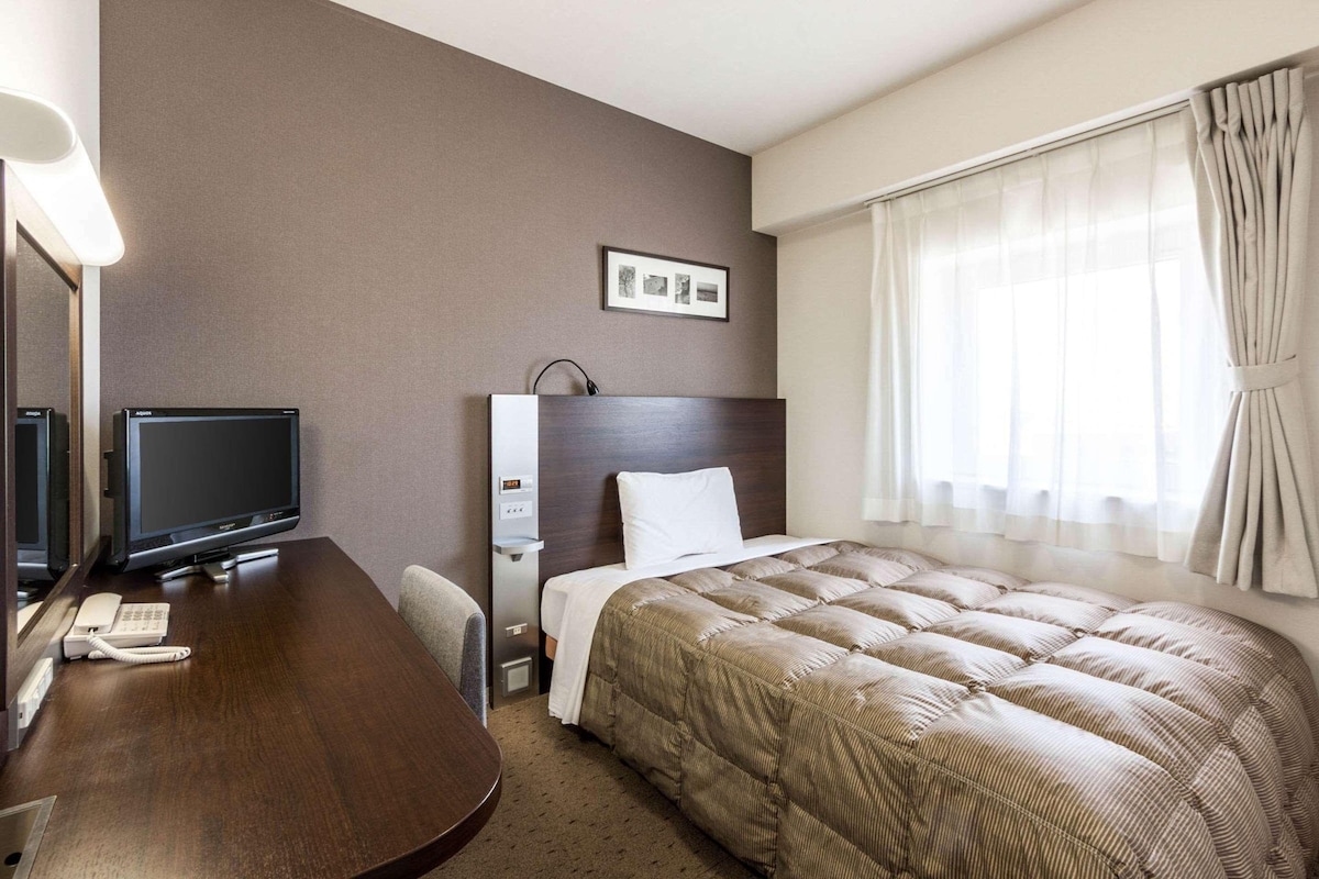 Una habitación de hotel con cama, escritorio con televisor, teléfono y un cuadro en la pared.