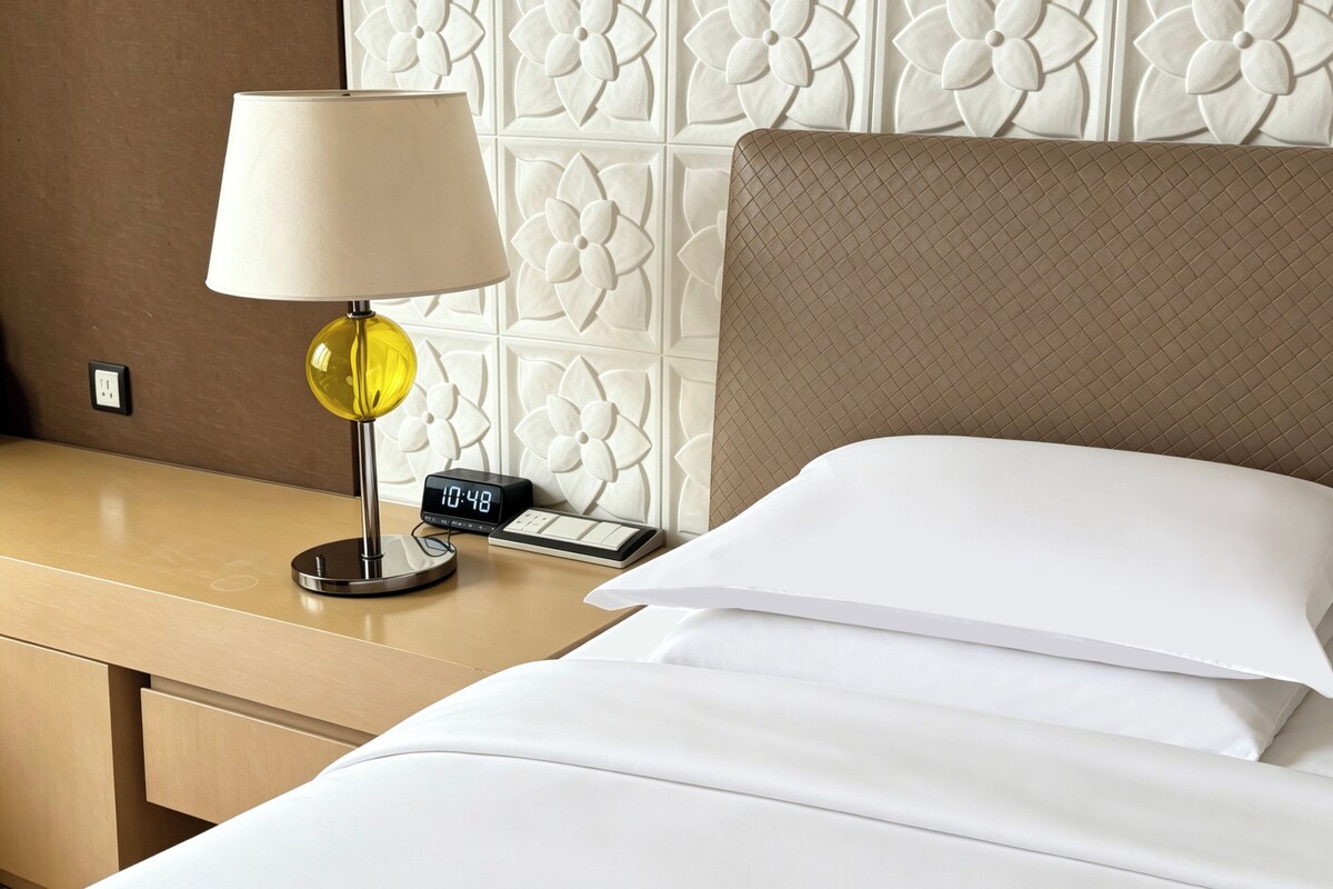 A hotel room with a bed, a nightstand, a lamp, a digital clock, and a wall with a floral pattern.