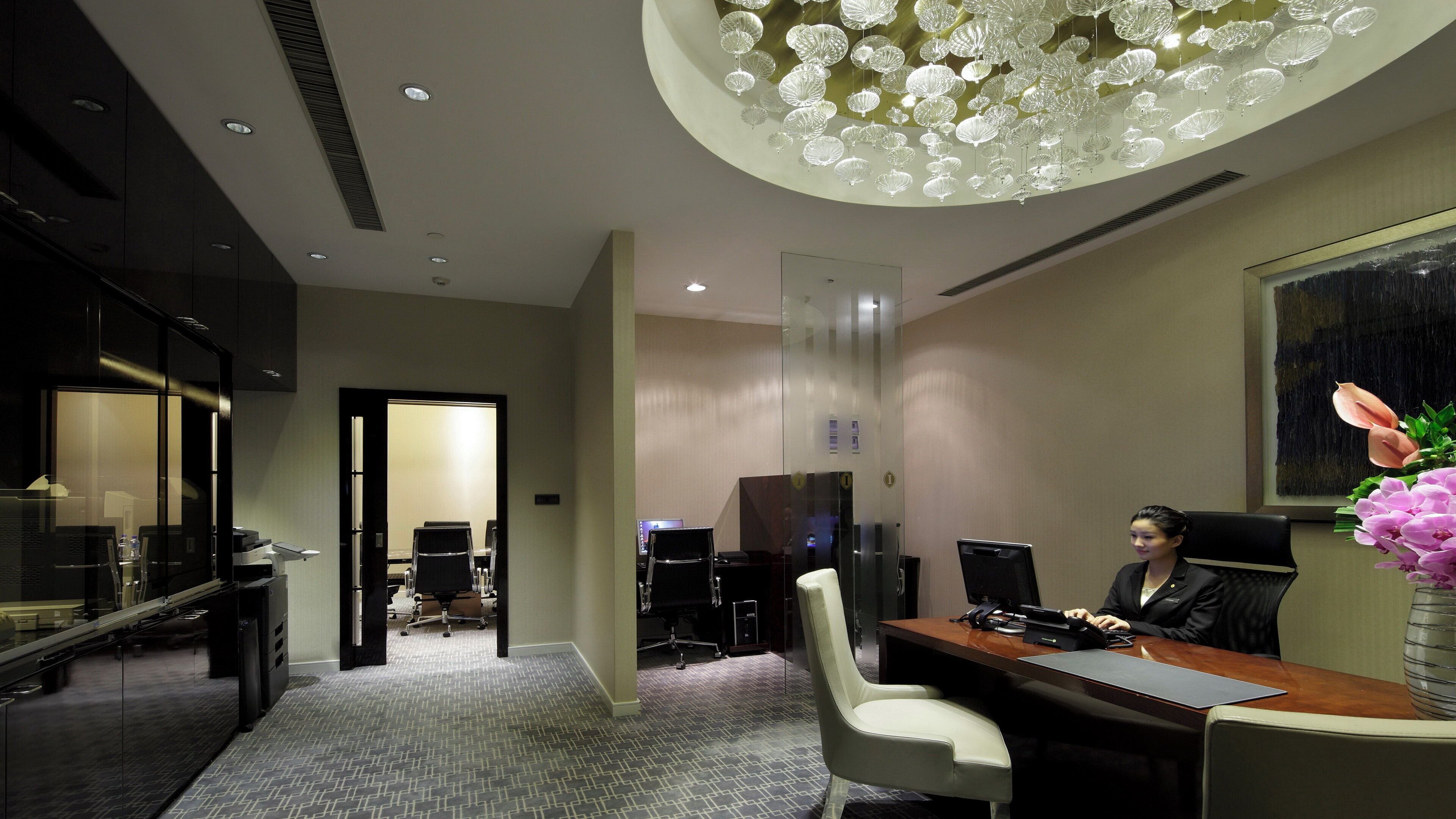 A modern office space with a desk, chairs, a large chandelier, and a painting on the wall.