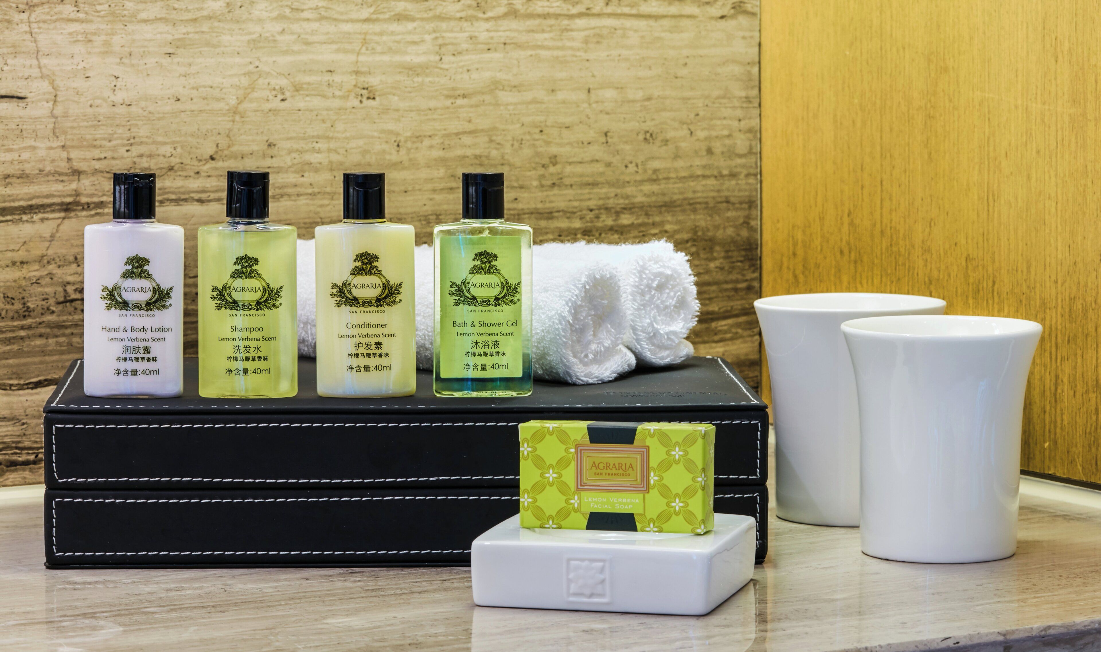 Conjunto de produtos de higiene pessoal sobre uma caixa preta com costura branca, uma toalha branca e duas chávenas de cerâmica brancas numa superfície de mármore.