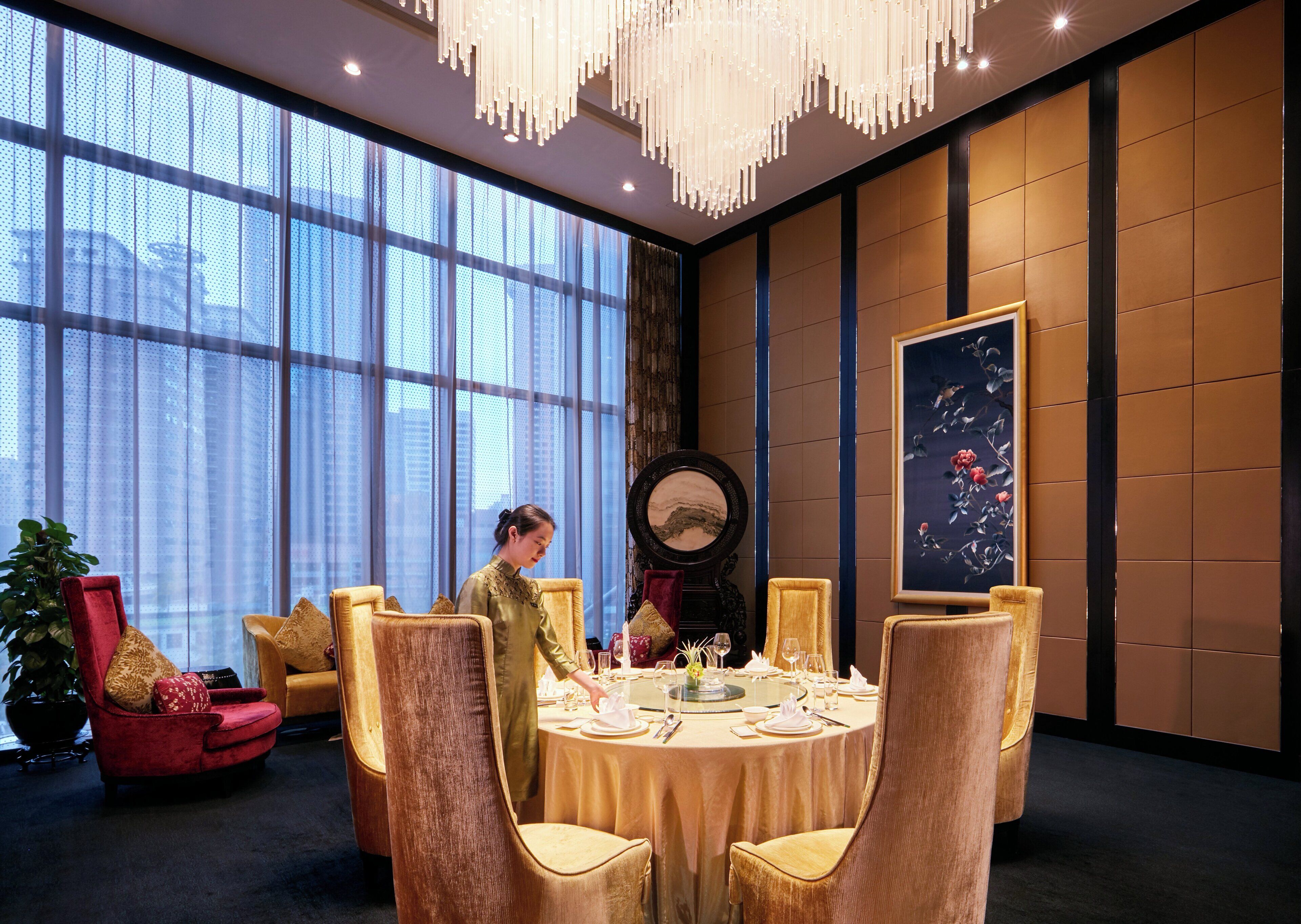 A dining area with a round table set for four, a large window with curtains, and a painting on the wall.