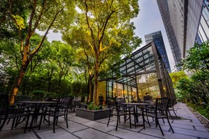 An outdoor cafe with tables and chairs, surrounded by trees and a modern building in the background.