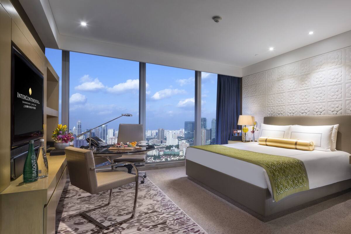 A modern hotel room with a large bed, a desk with a computer, a chair, a TV, and a city view.