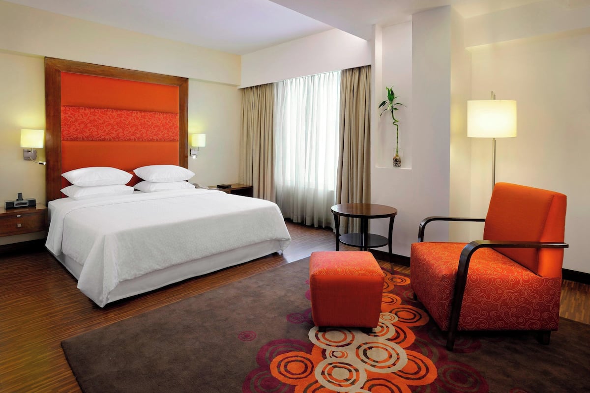 A hotel room with a bed, a chair, a small table, a lamp, and a window with curtains.