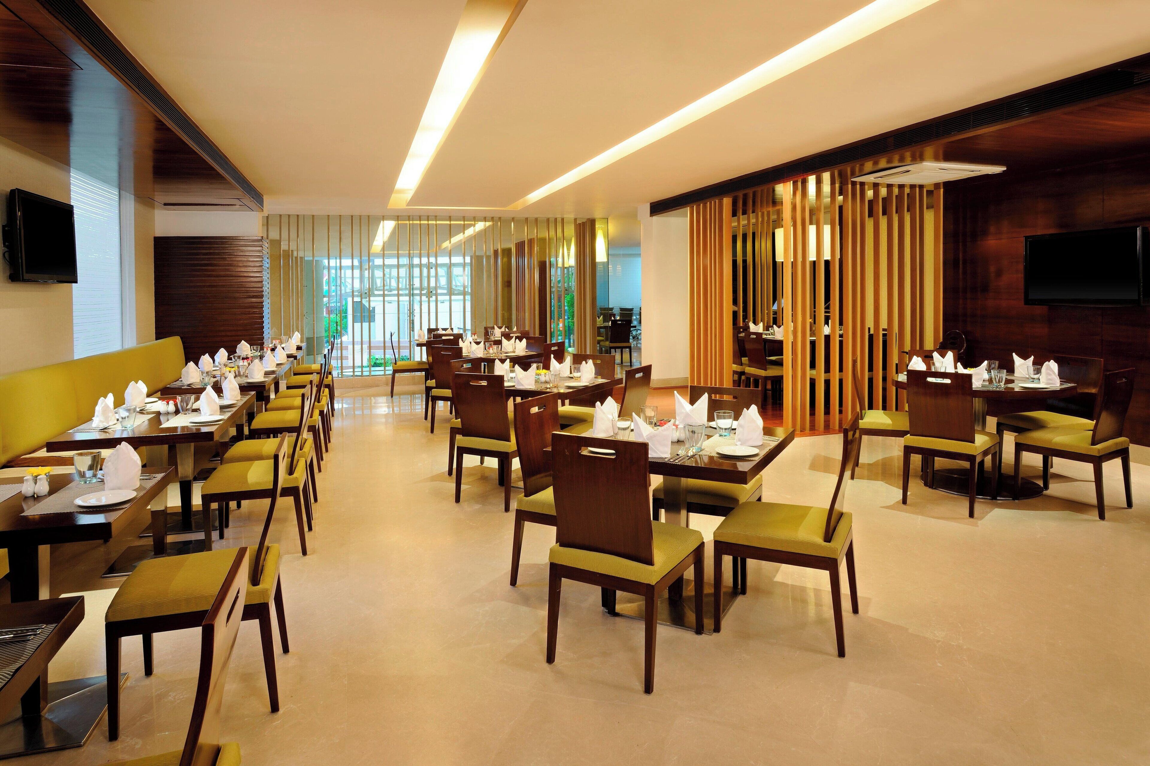 A spacious dining area with multiple tables and chairs, a television mounted on the wall, and large windows allowing natural light.