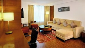 A hotel room with a sofa, armchair, coffee table, and a desk with a lamp.