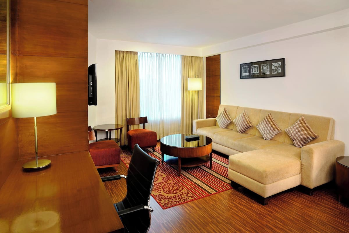 A hotel room with a sofa, armchair, coffee table, and a desk with a lamp.