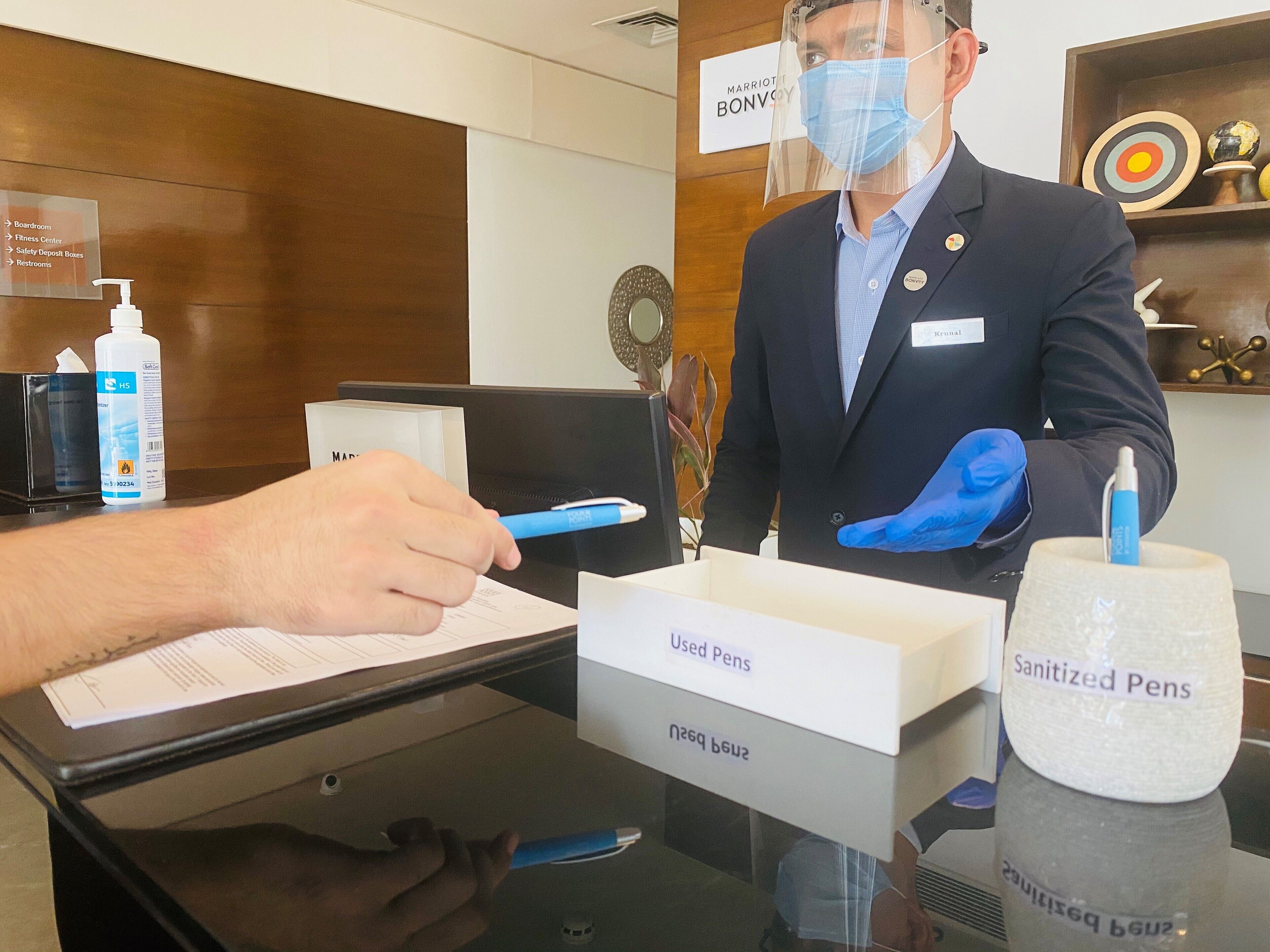 Un mostrador de recepción de hotel con un cartel que indica servicios como una sala de juntas, gimnasio y baños. Un huésped está firmando un documento, y hay cajas etiquetadas como 'Bolígrafos Usados' y 'Bolígrafos Sanitizados'.