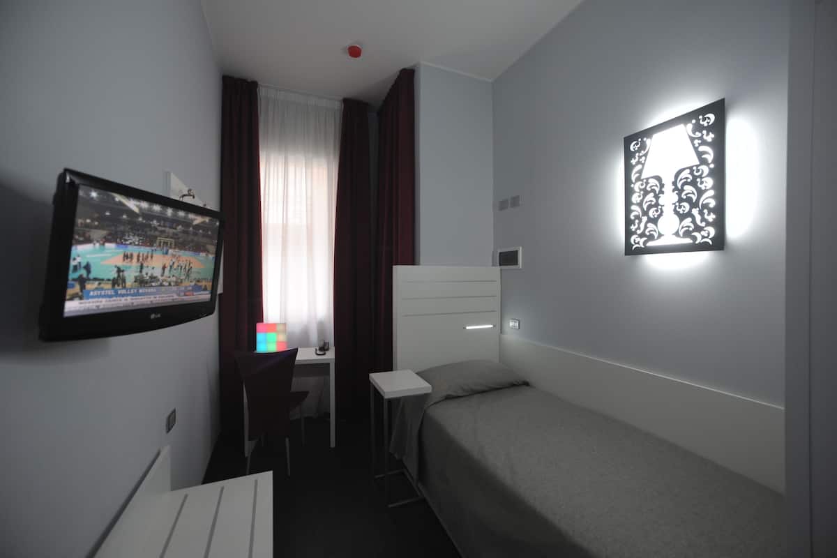 A hotel room with a bed, a desk, a chair, a TV, and a wall-mounted light fixture.
