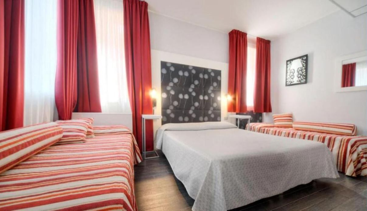 Een hotelkamer met twee bedden, rode gordijnen, een kunstwerk aan de muur en een nachtkastje met een lamp.