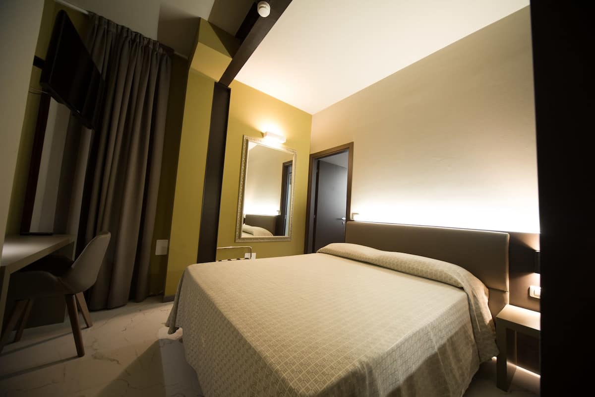 A hotel room with a bed, a desk, a chair, a mirror, and a door.