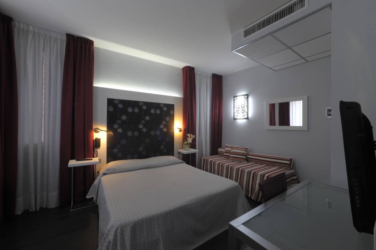 A hotel room with a bed, a bedside table, a lamp, a mirror, and a window with curtains.