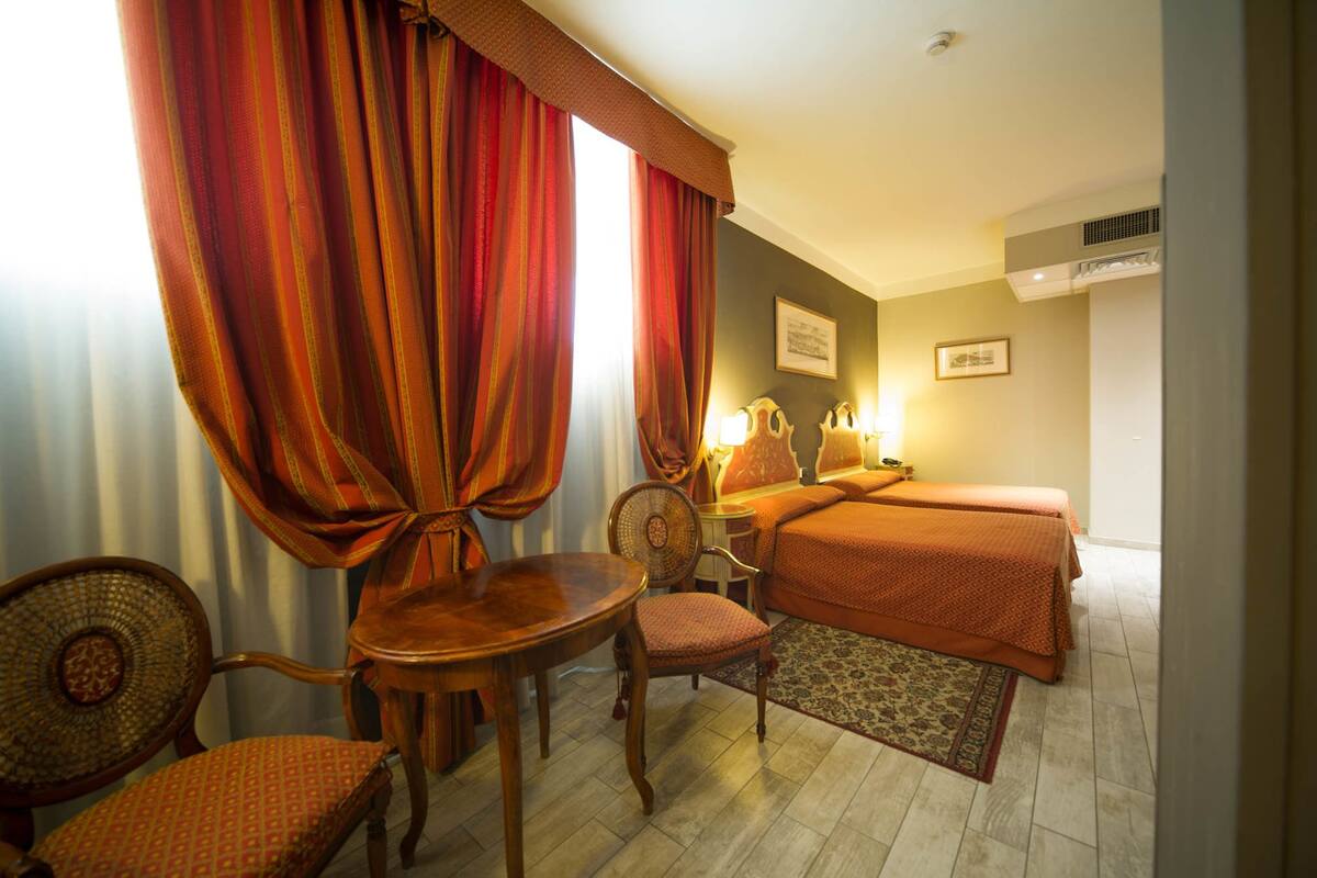 A hotel room with two beds, a dining table with chairs, a small table with a chair, and a large window with curtains.