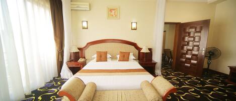 A hotel room with a large bed, two bedside tables, a chair, a standing fan, and a door leading to another room.