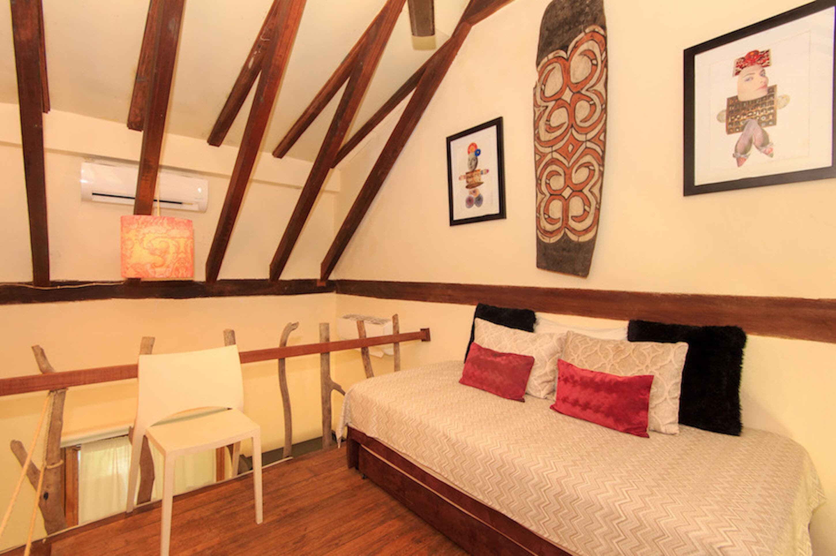 Une chambre avec un plafond en bois, un lit recouvert d’une literie à motifs, une chaise et des œuvres d’art accrochées aux murs.