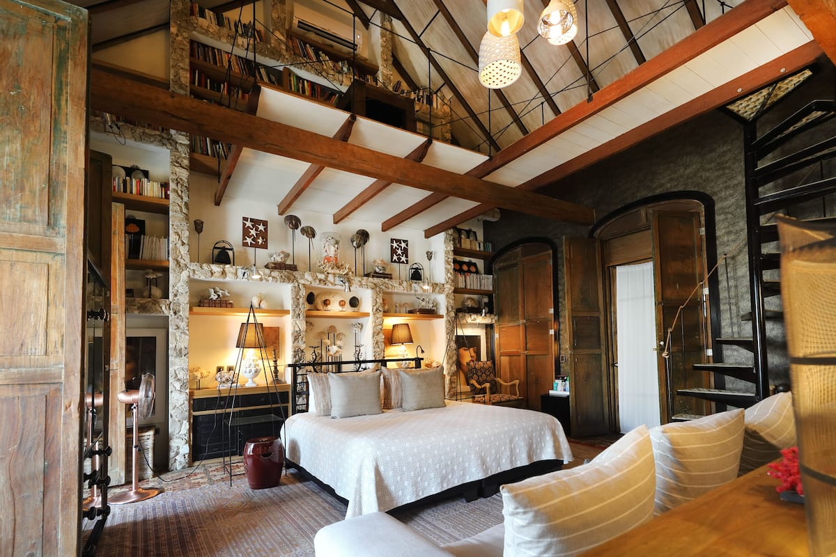 A spacious bedroom with a large bed, wooden beams, stone walls, and a high ceiling.