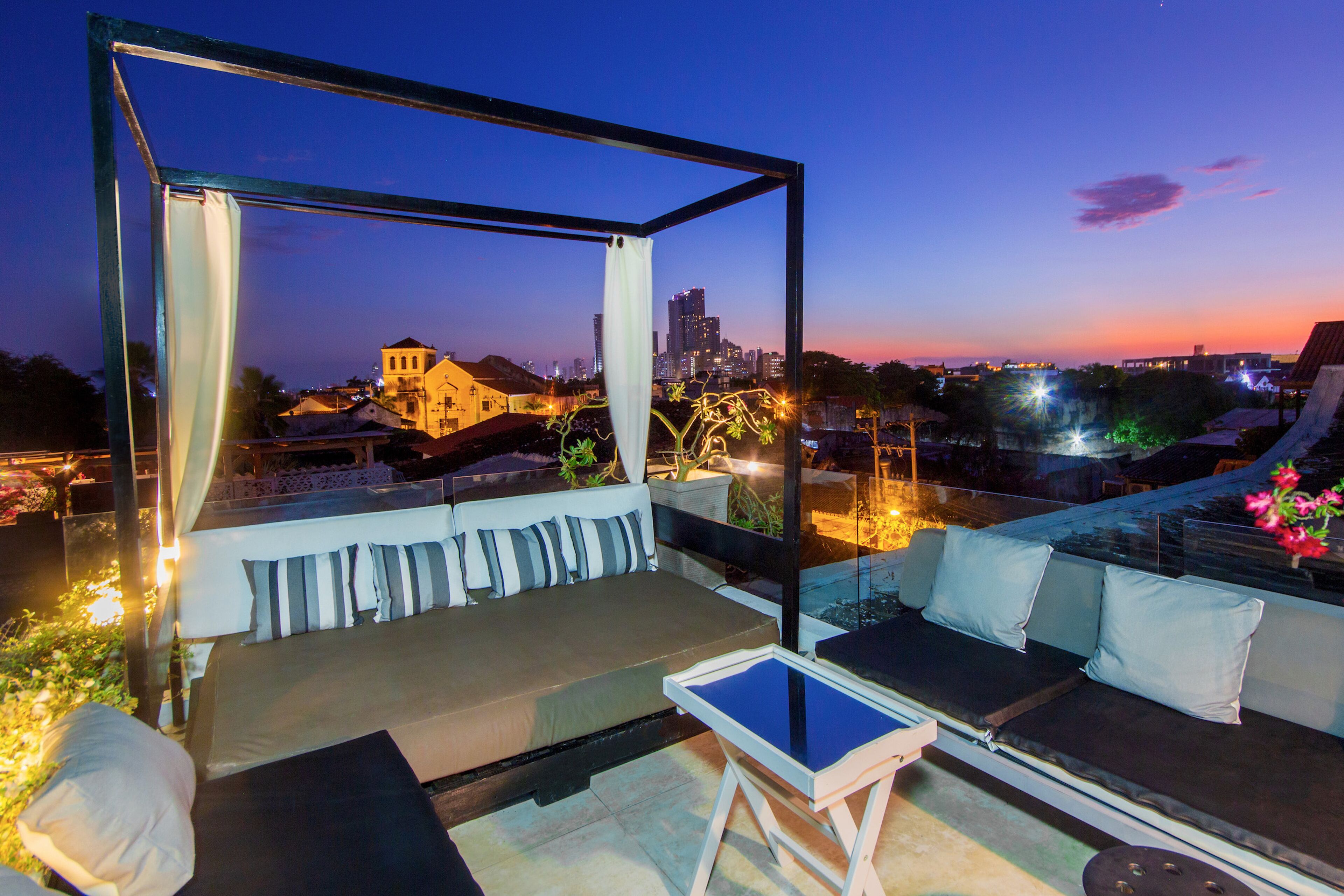 A rooftop terrace with a view of a city skyline at dusk, featuring a seating area with cushions and a small table.