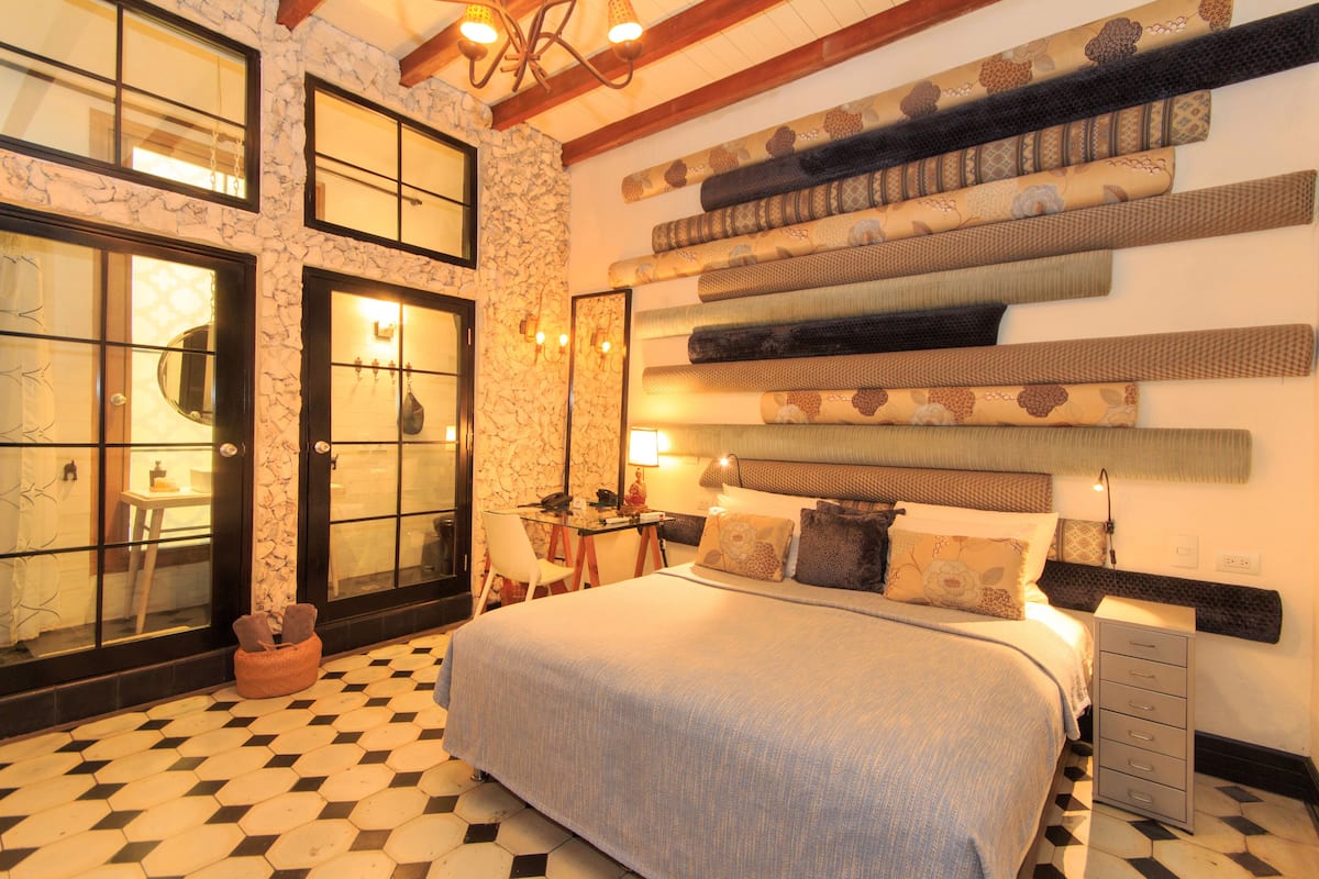 A hotel room with a bed, a desk, and a chair. There is a wall with patterned fabric hanging above the bed.