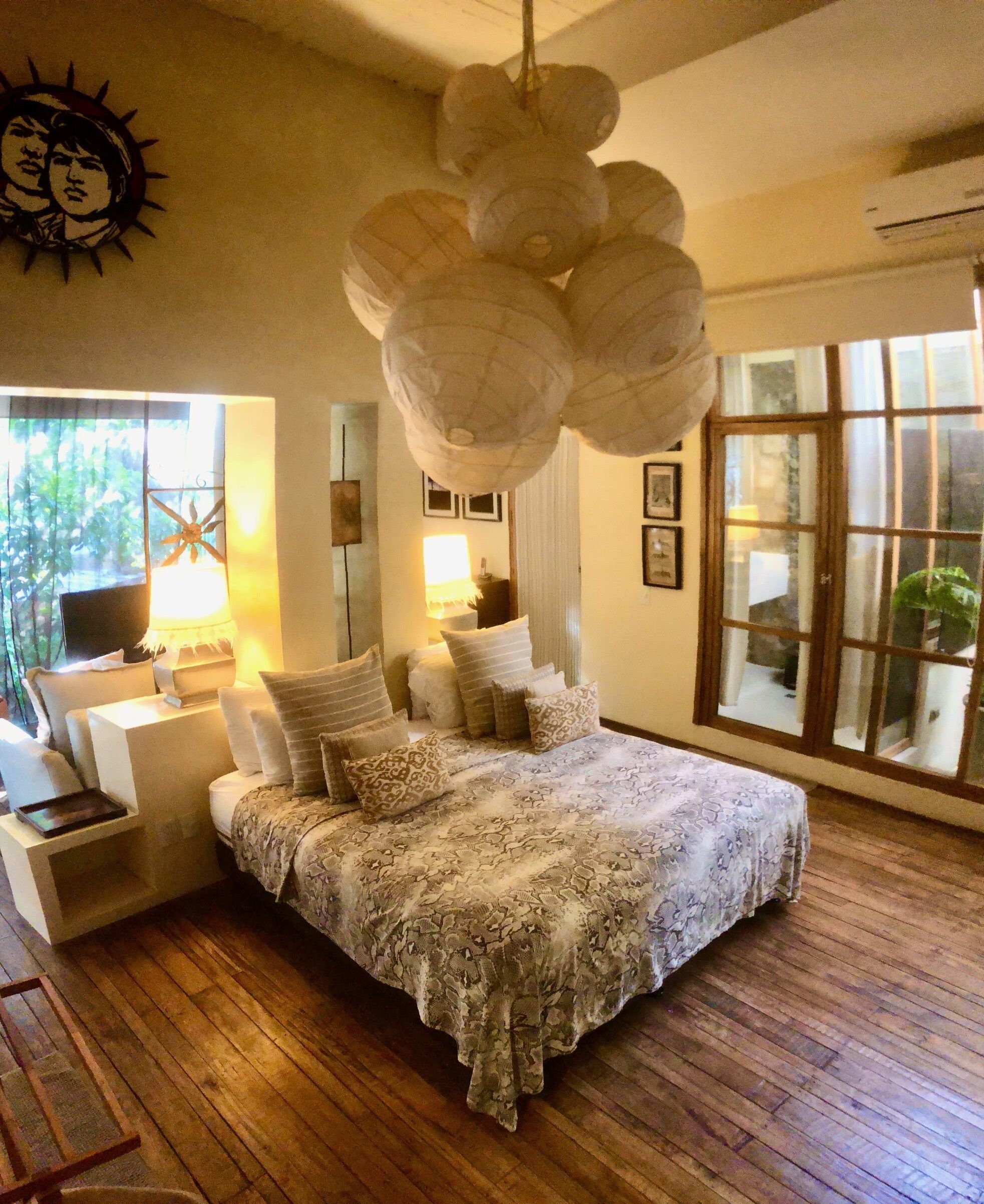 A bedroom with a large bed, a desk, and a chair. There are multiple pillows and a hanging light fixture.
