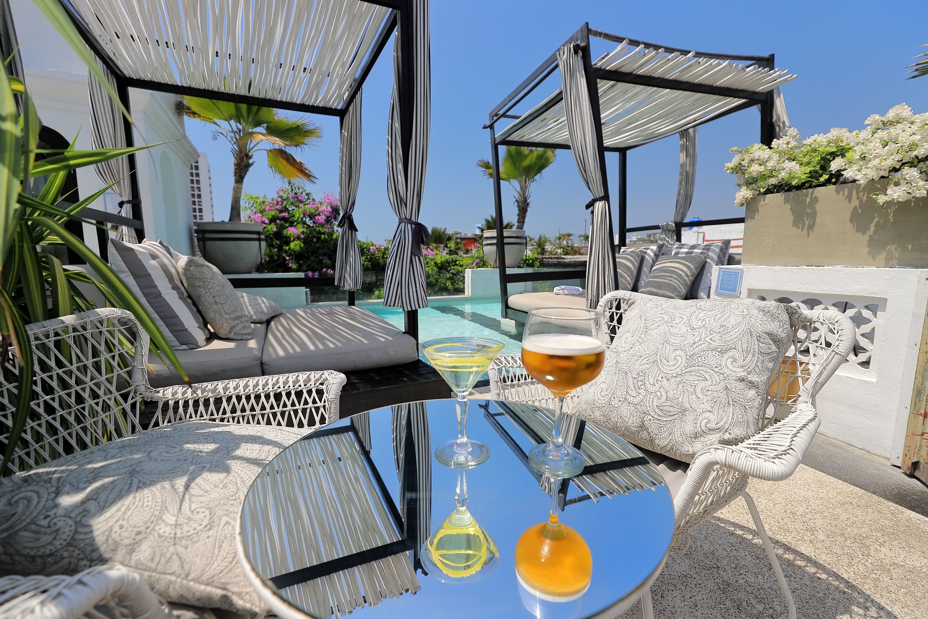 Un área de lounge en la azotea con mobiliario exterior, piscina y una mesa de vidrio con bebidas.