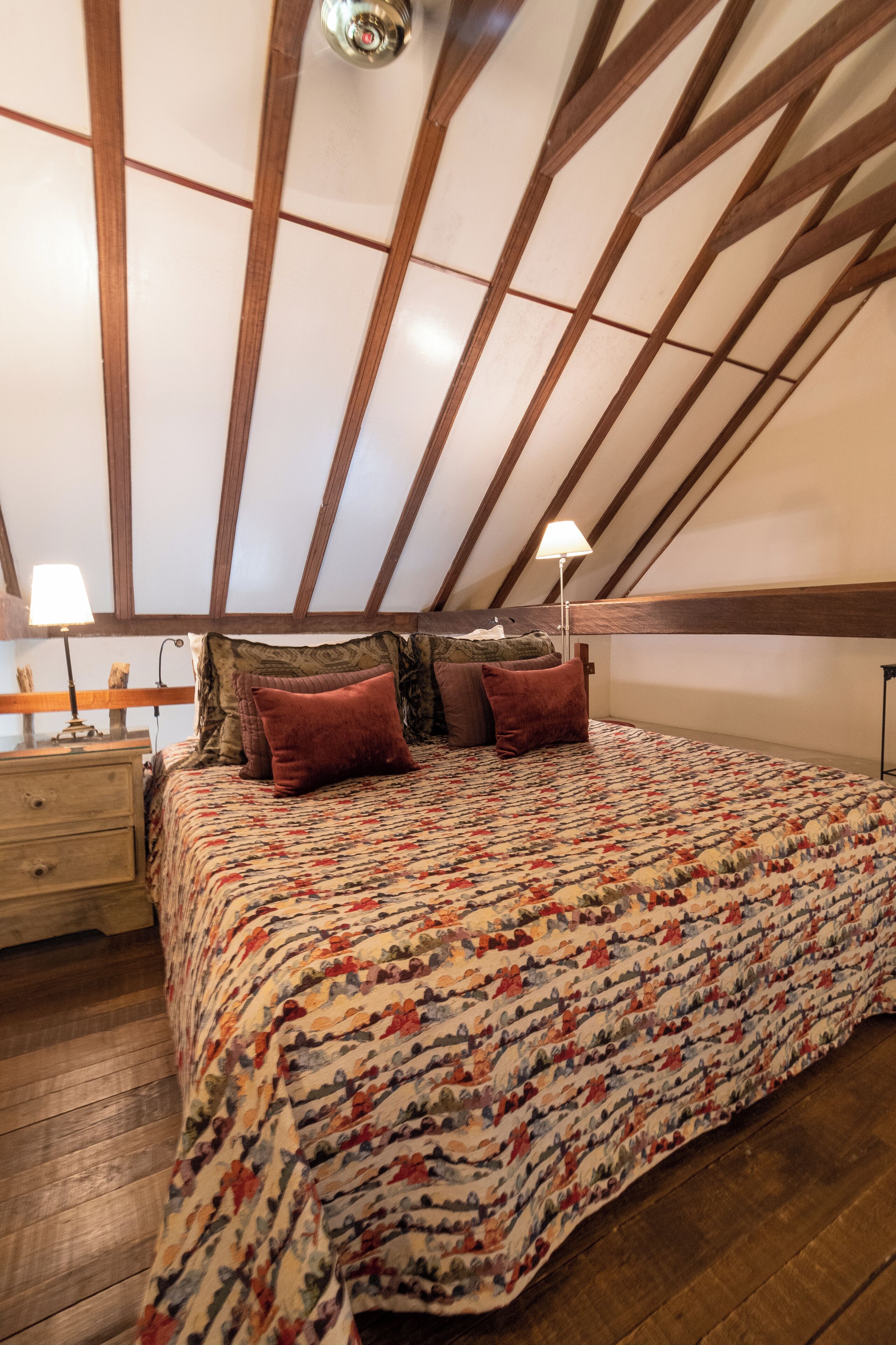 A bedroom with a sloped ceiling, wooden beams, a bed with a patterned bedspread, two bedside lamps, and a nightstand.