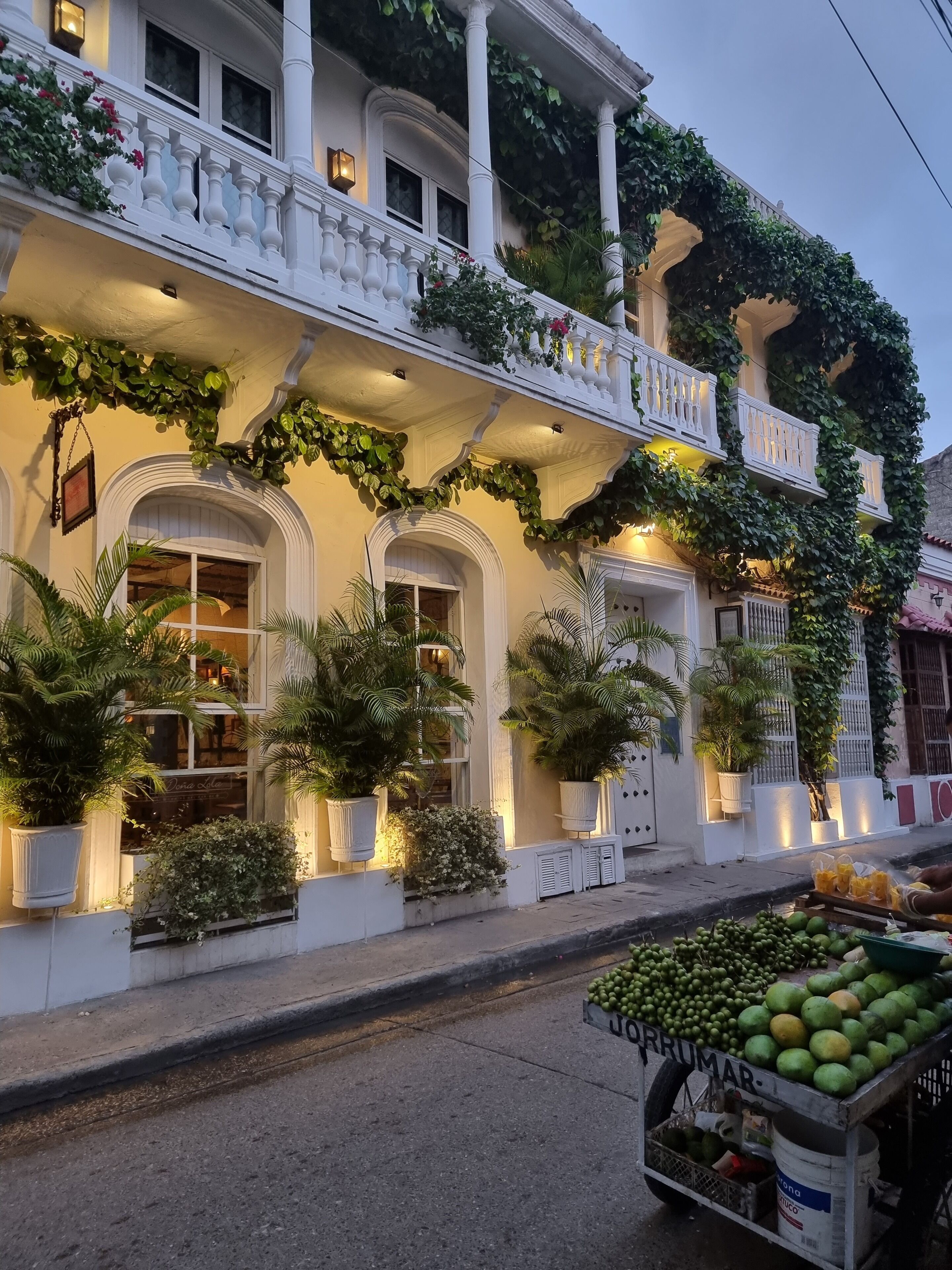 A building with white exterior walls, multiple balconies with white railings, and plants. There is a fruit cart on the street in front.