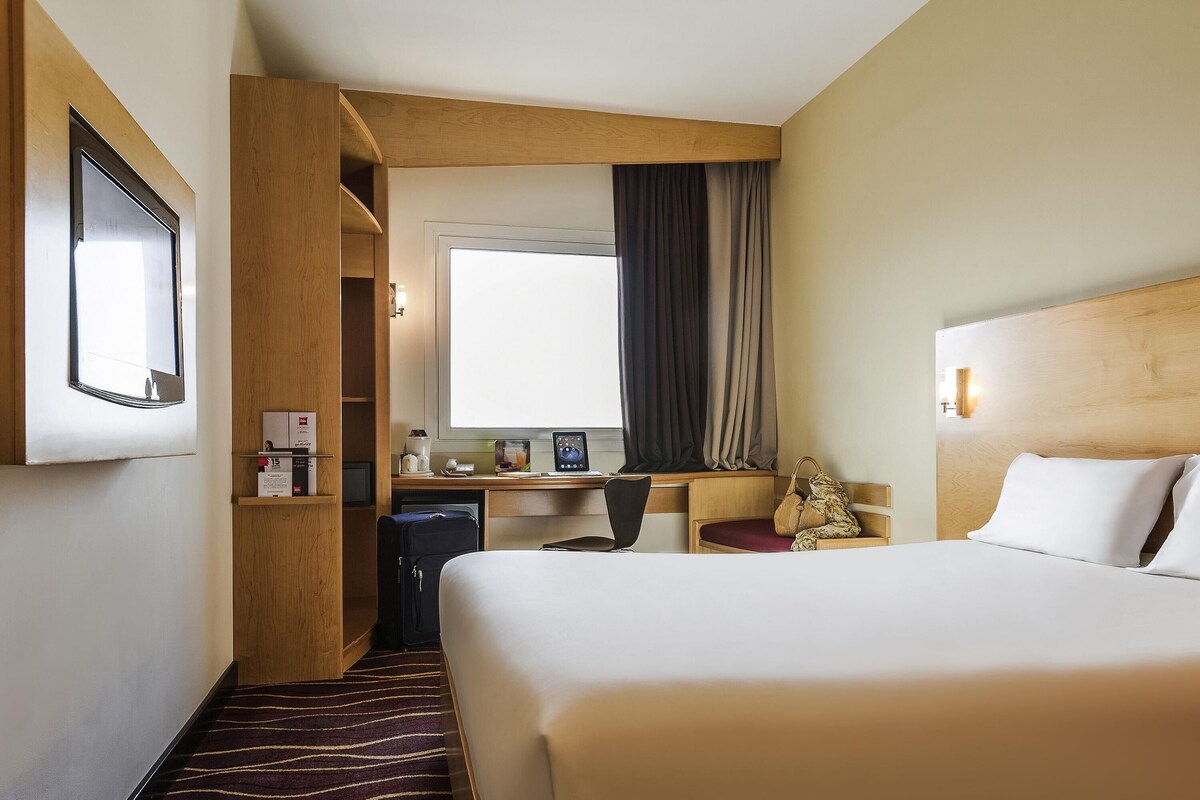 A hotel room with a large bed, a desk with a computer, a chair, a small table with a lamp, a window with curtains, and a wall-mounted TV.