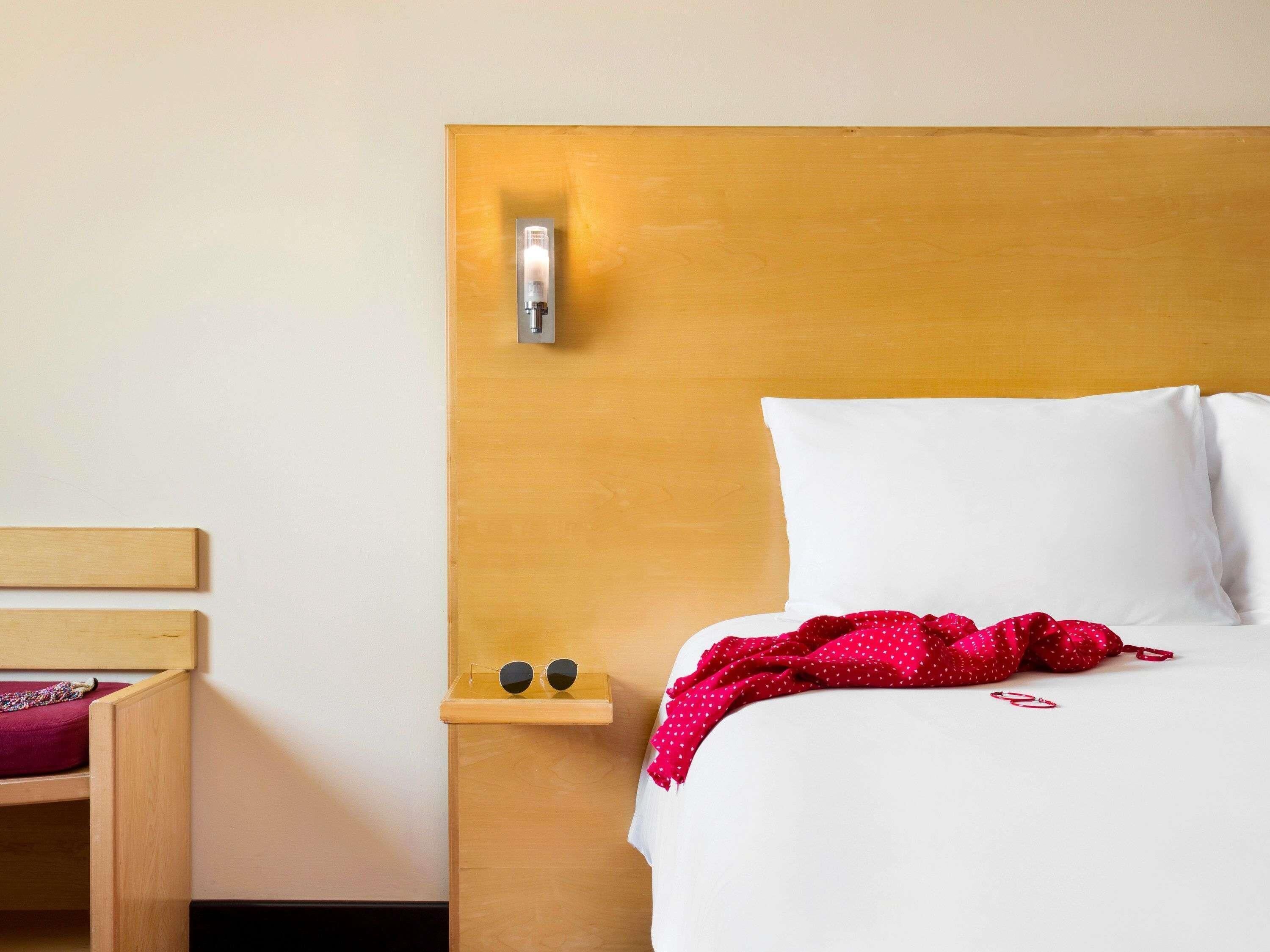 A hotel room with a wooden headboard, a bedside table with a lamp, a small shelf with sunglasses, and a bed with white bedding and a red polka dot blanket.