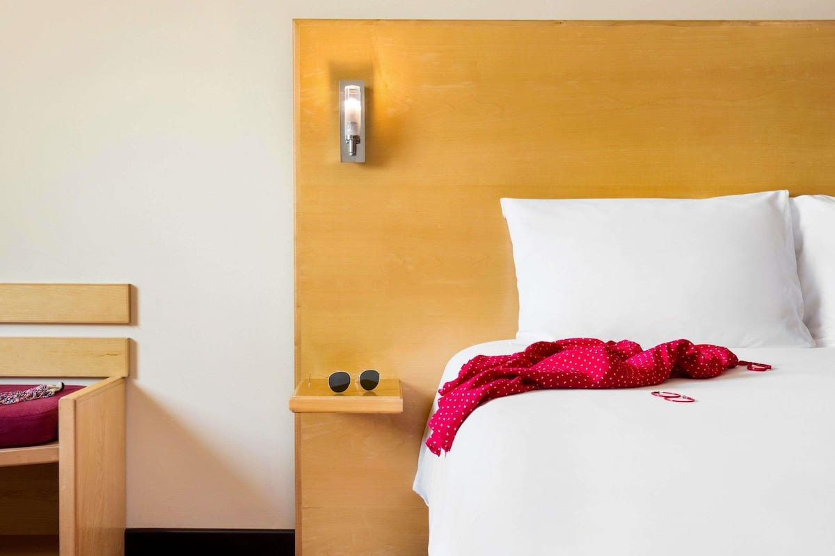 A hotel room with a wooden headboard, a bedside table with a lamp, a small shelf with sunglasses, and a bed with white bedding and a red polka dot blanket.