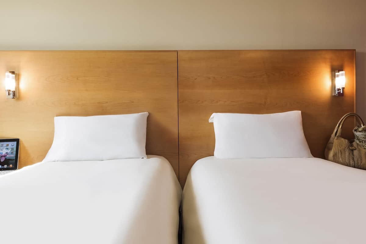 Habitación de hotel con dos camas, cada una con sábanas blancas y cabeceros de madera. Hay luces fijadas a la pared a cada lado del cabecero. Se ve una tableta en el lado izquierdo de la cama.
