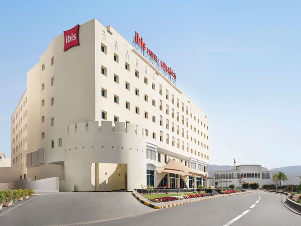 A hotel with a prominent 'ibis Hotel' sign, a large entrance with a covered area, and a landscaped garden in front.