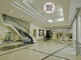 Egy modern szállodai előcsarnok, központi lépcsőházzal, pihenőhelyekkel és feltűnő „IBIS HOTEL” felirattal.