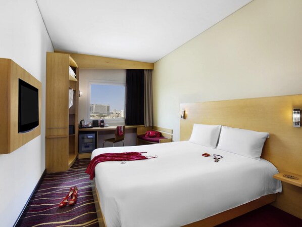 A hotel room with a bed, a desk, a television, and a view of the city. - ibis Muscat (Muscat)