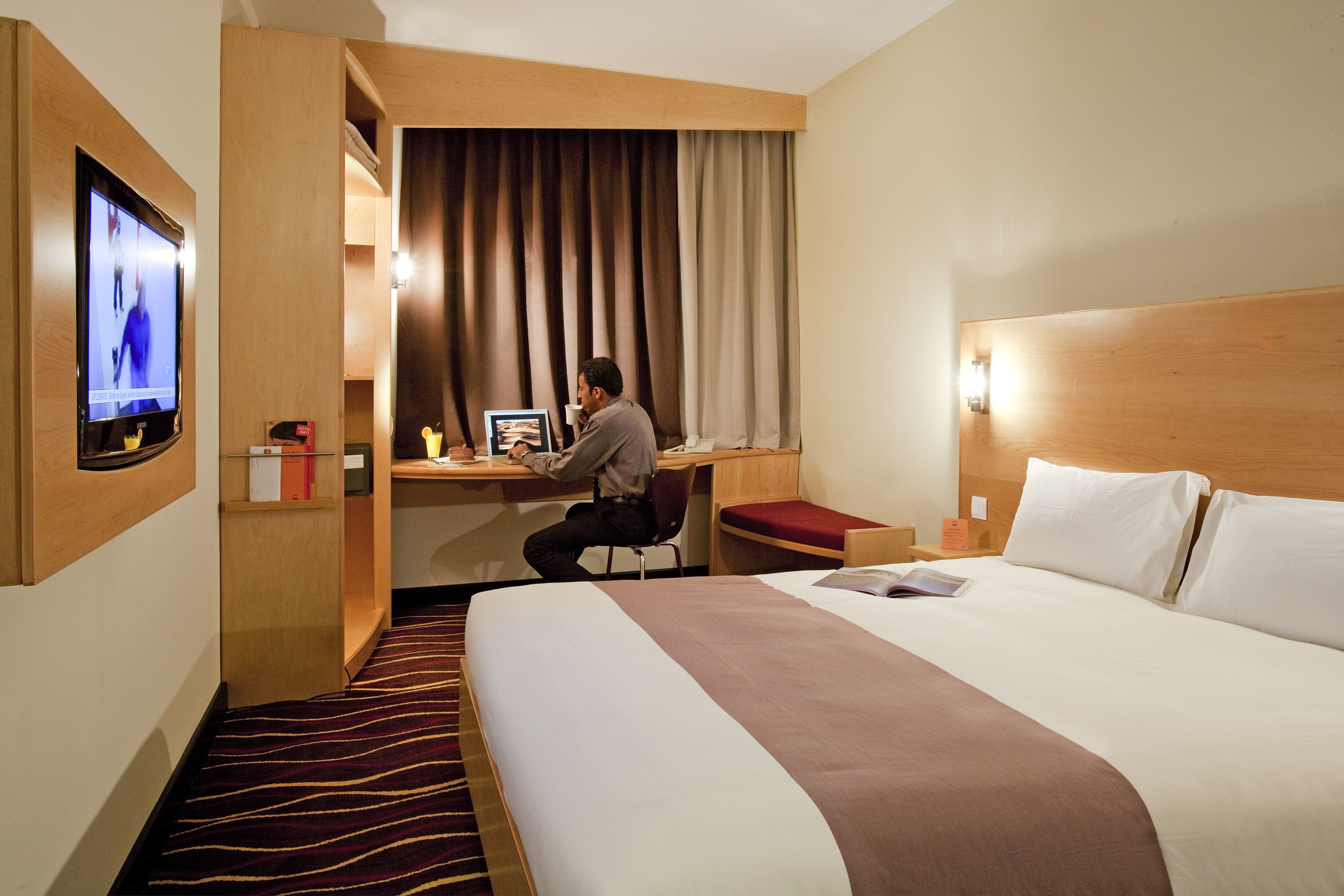 Quarto de hotel com cama, escrivaninha, cadeira, televisão e janela com cortinas.