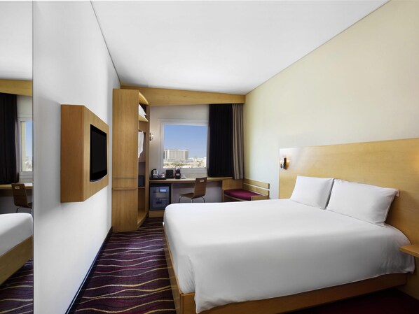 A hotel room with a bed, desk, chair, TV, and a view of the city. - ibis Muscat (Muscat)