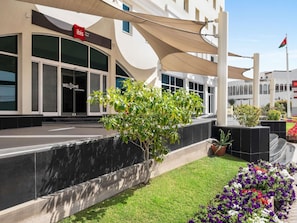 A modern hotel entrance with a 'Welcome to My World' sign, a covered outdoor area with plants, and a flagpole with a flag. - ibis Muscat (Muscat)