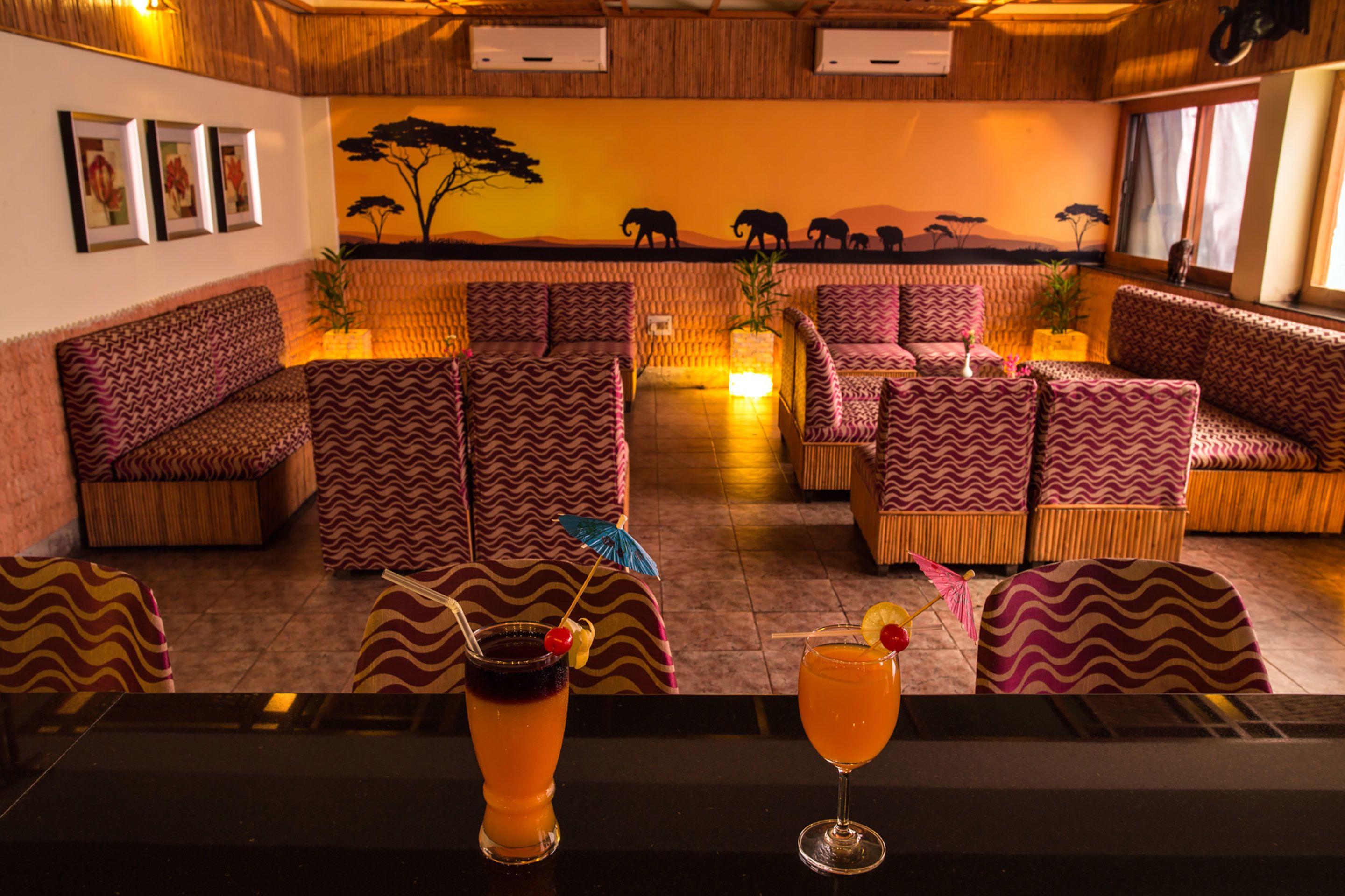 A lounge area with patterned sofas and chairs, a bar counter with drinks, and a mural of elephants on the wall.
