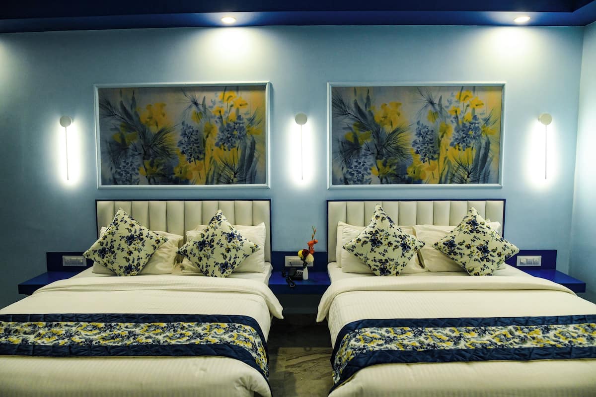 Habitación de hotel con dos camas, cada una con una colcha de estampado floral, una mesita de noche con lámpara, y dos cuadros de flores colgados en la pared.