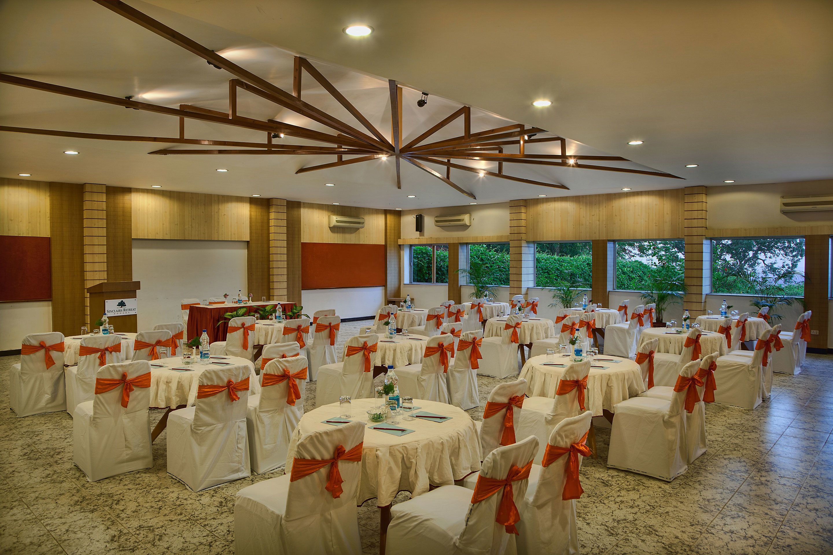 Un salón de banquetes con mesas redondas, sillas con cintas naranjas y un diseño moderno en el techo.