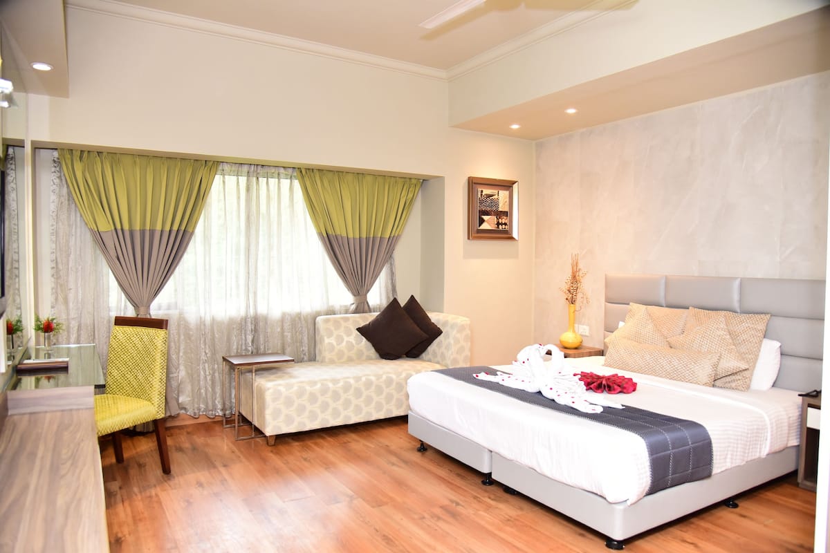 Habitación de hotel con cama, sofá, sillón, mesita de noche y ventana con cortinas.