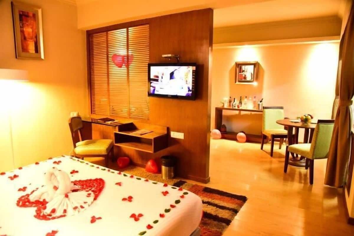 Una habitación de hotel con una cama decorada con pétalos de rosa, un televisor en la pared, una zona de estar con una mesa y sillas, y una kitchenette pequeña.