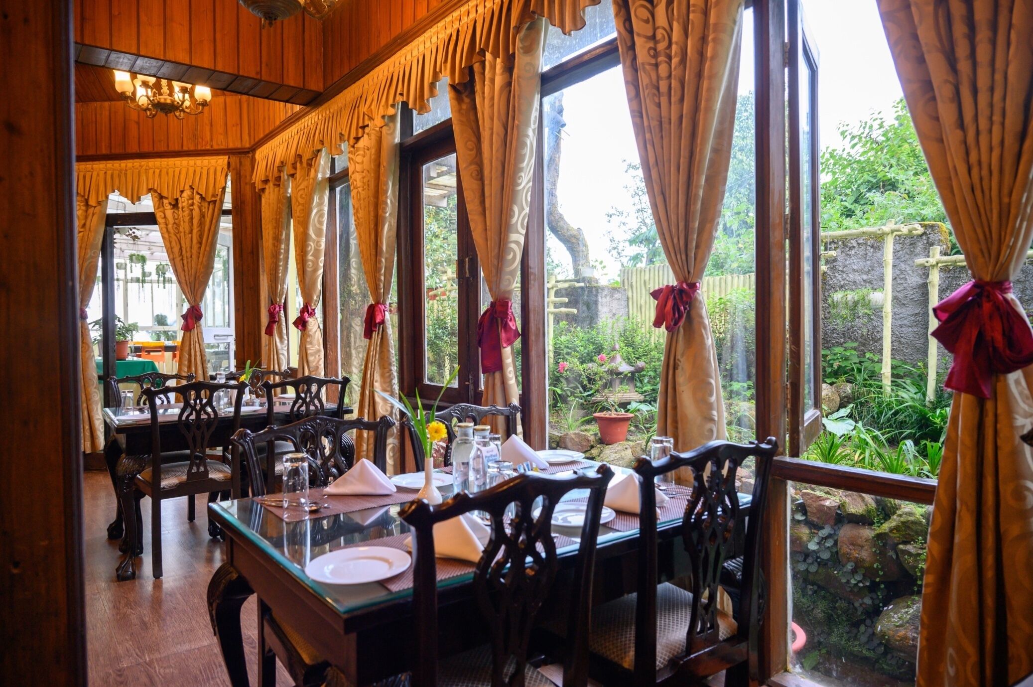 A dining area with wooden furniture, large windows with curtains, and a view of greenery outside.
