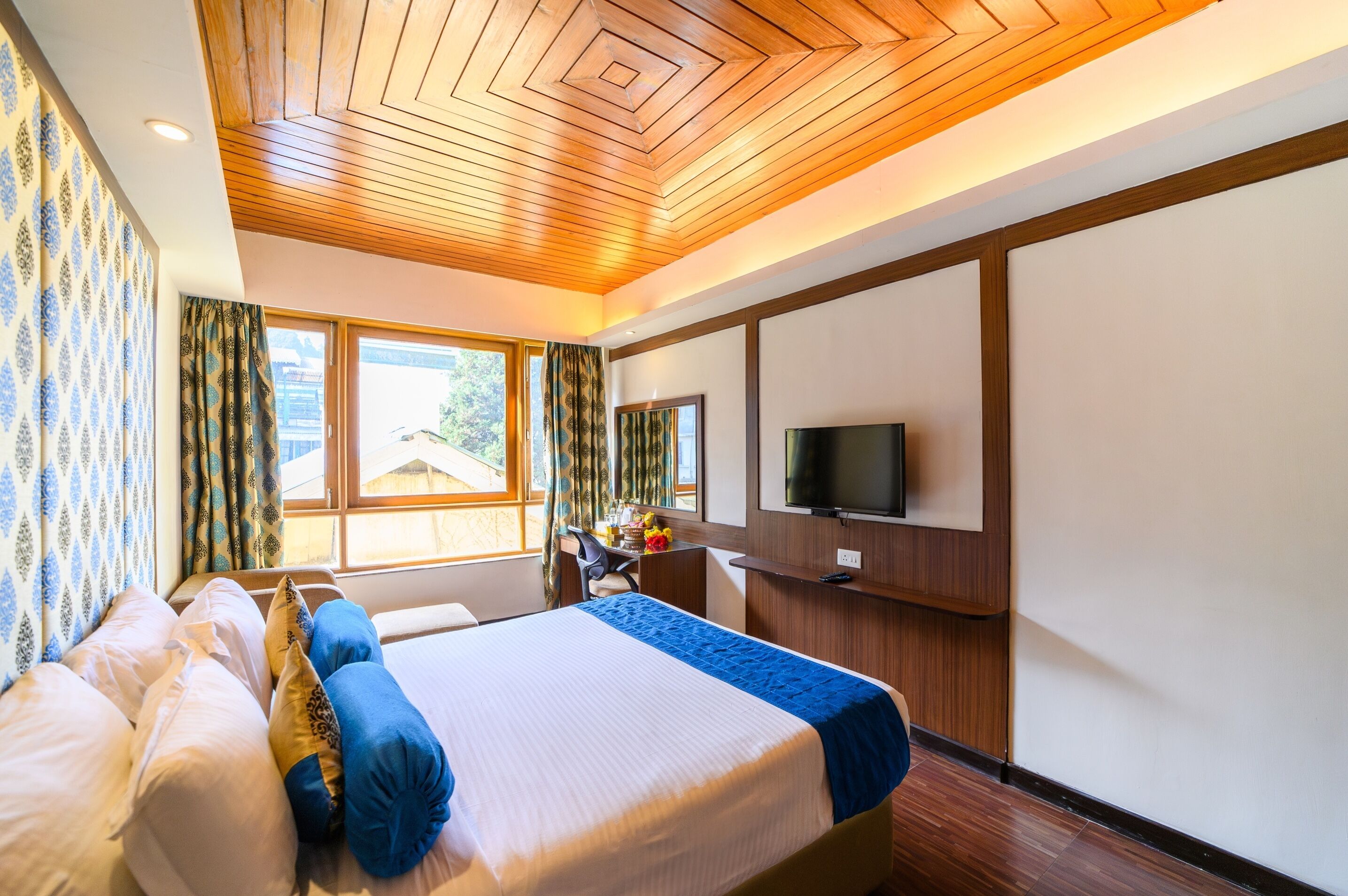 A hotel room with a large bed, a desk with a chair, a TV mounted on the wall, and a window with curtains.