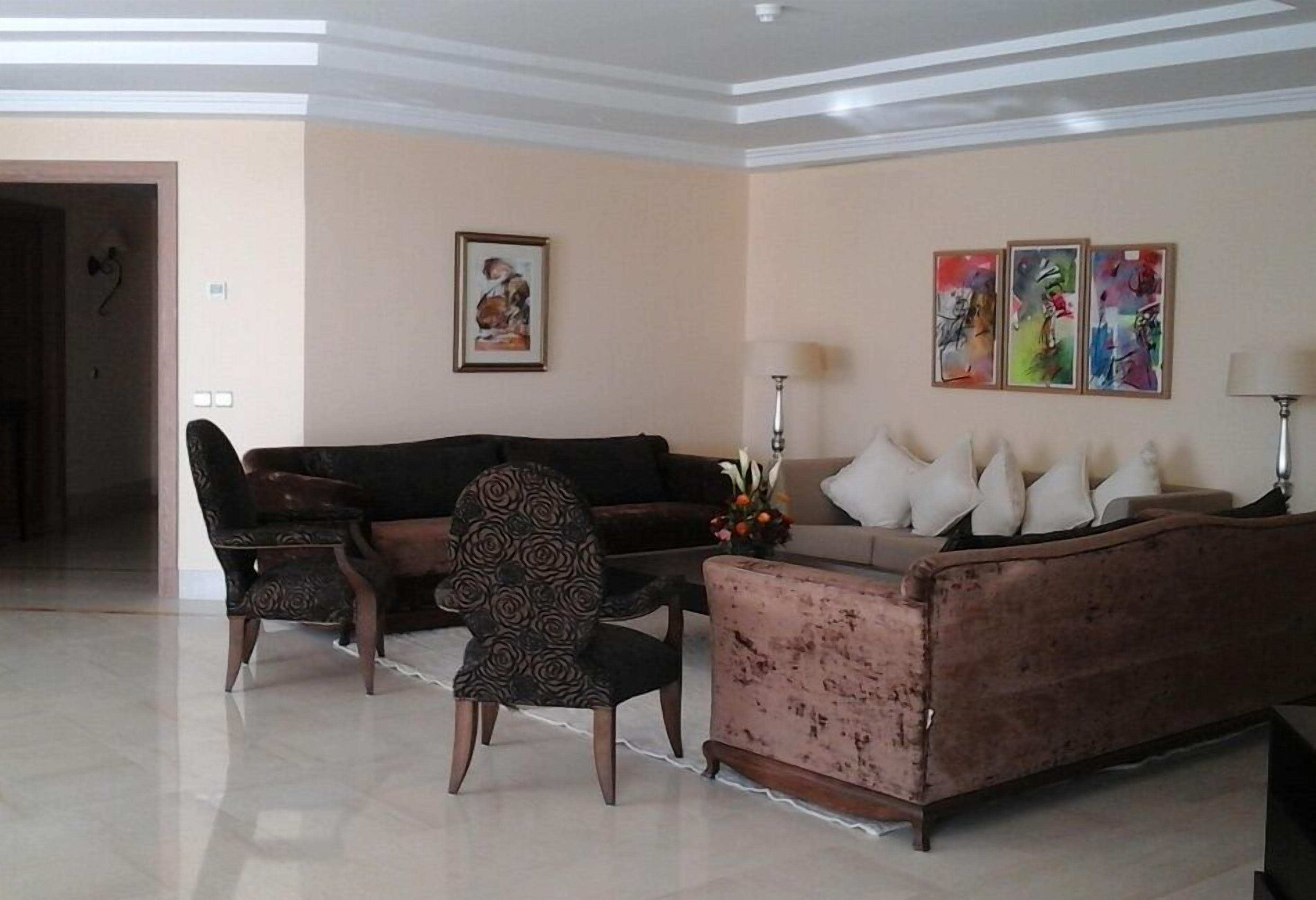 A living room with a sofa, armchairs, and a coffee table. There are wall-mounted lamps and a framed picture on the wall.