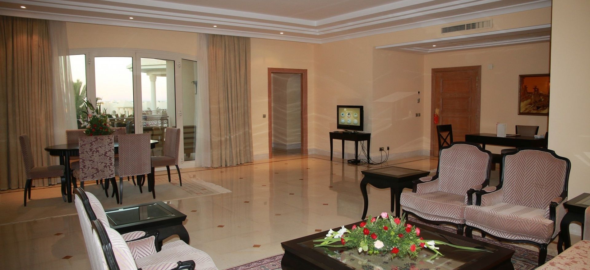 A spacious living room with a glass door leading to a balcony, a television, and a coffee table with flowers.