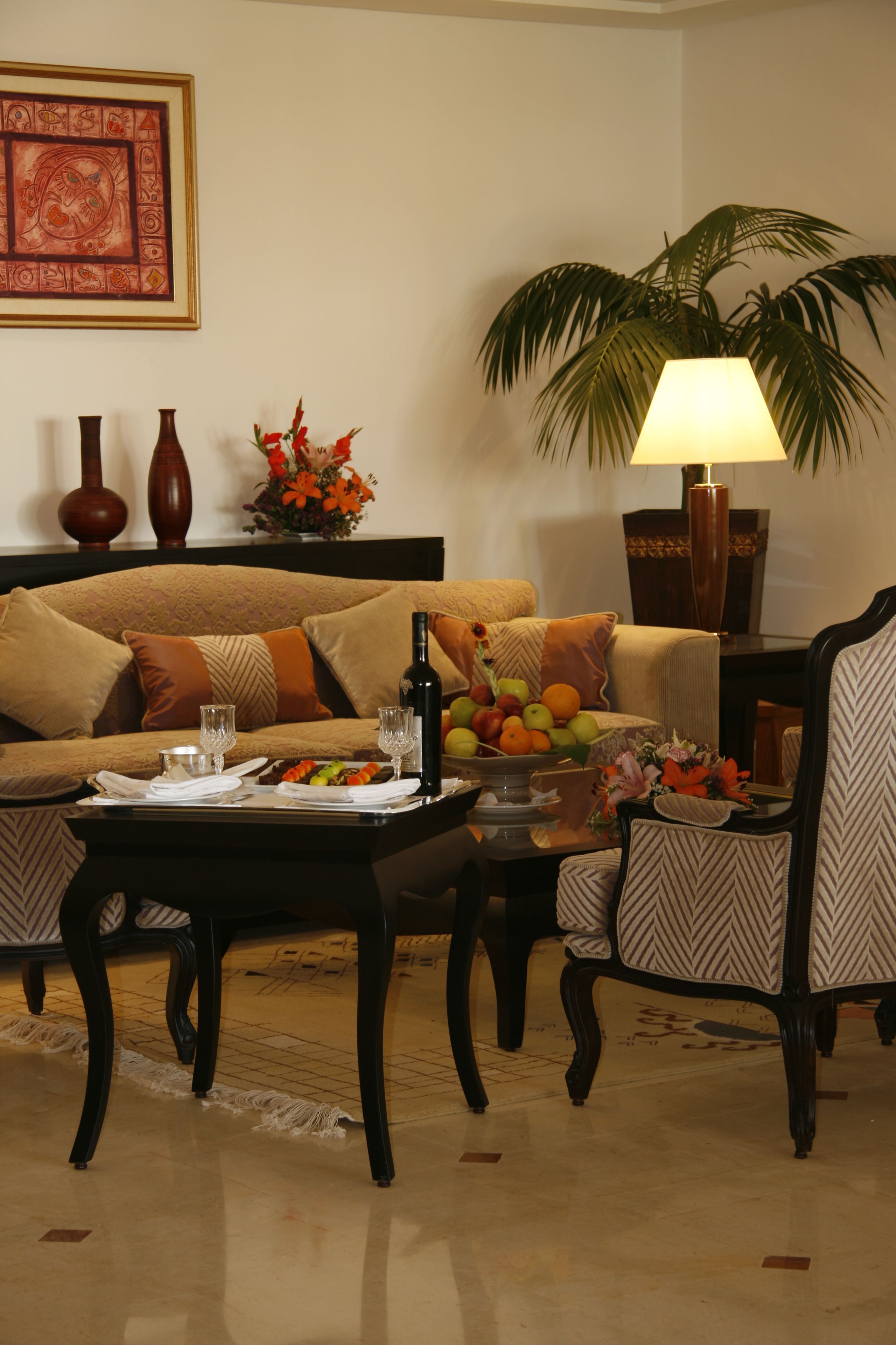 A living room with a sofa, coffee table, and two chairs. There is a lamp, a vase with flowers, and a fruit bowl on the table.